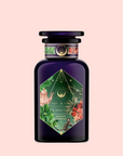 A dark purple perfume bottle with a hexagonal floral label of green leaves and pink flowers sits on a pale pink background. The label reads "Moonlight in Marrakesh: Spiced Mint Green Tea.