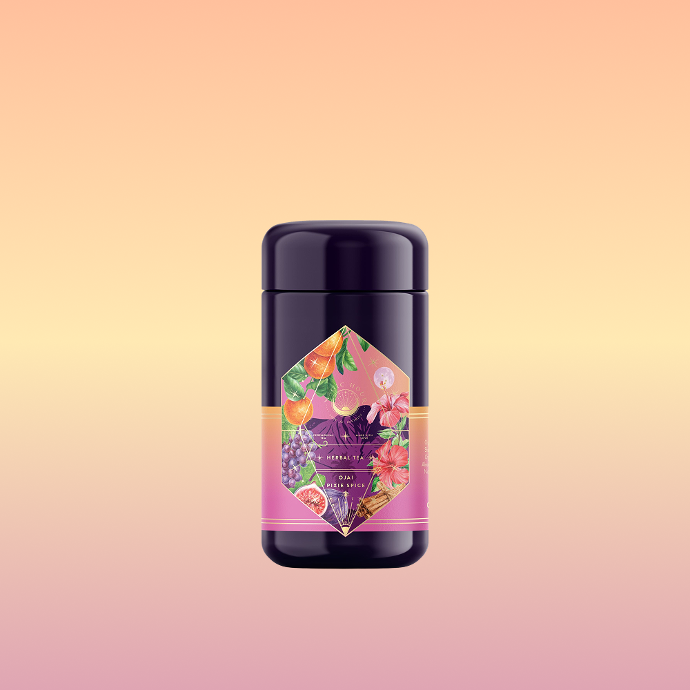 A dark purple cylindrical container of Pink Moment Pixie - Hibiscus Tangerine Dream Herbal Tea, with a colorful label showing oranges, grapes, flowers, and elderberry, set on a soft peach and yellow gradient background.
