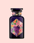 The Pink Moment Pixie - Hibiscus Tangerine Dream Herbal Tea comes in a dark purple glass bottle with a black cap and a vibrant label featuring grapes, oranges, and pink flowers on a light pink background.