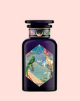 The Tulum Horchata Chai perfume comes in a dark purple bottle with a colorful, geometric label showing tropical birds, flowers, and a white-clad figure. Inspired by adaptogenic potions, it features a round cap and ornate details against a pale pink background.