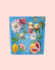 A vibrant tea package with botanical illustrations of flowers, lemons, and figs on a blue background, labeled "Summer in Amalfi" in gold—celebrating Mediterranean tea with organic lemon peel notes.