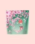 A Sencha Green Tea package called "Queen of Kyoto," decorated with pink cherry blossoms and hummingbirds on a teal background, set against a light pink backdrop—evoking the Japanese Cherry Blossom Festival.