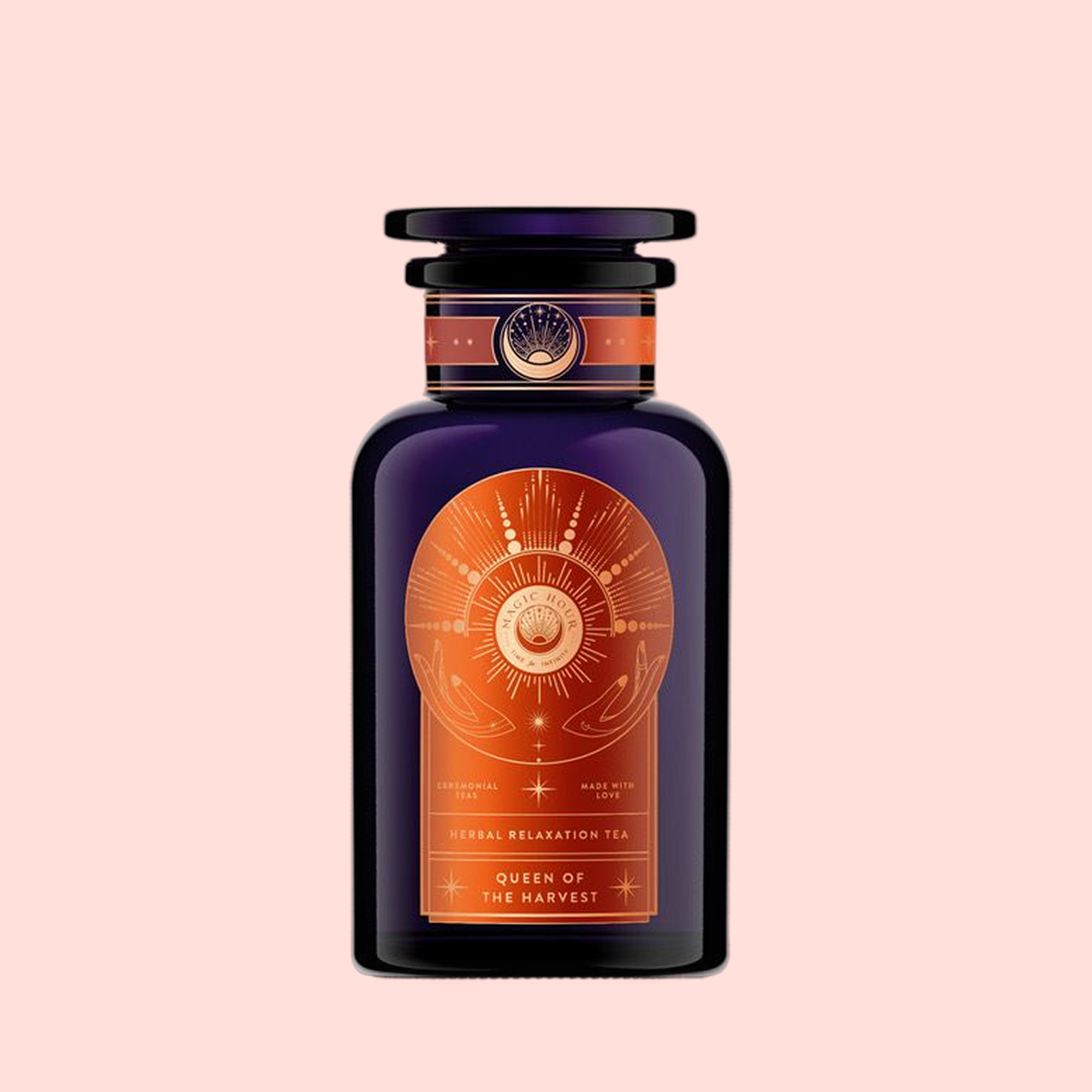 A dark purple glass bottle with a gold and copper celestial label reads “Queen of the Harvest: Herbal Relaxation Tea,” featuring organic herbal tea, set against a light pink background.