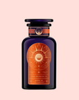 A dark purple glass bottle with a gold and copper celestial label reads “Queen of the Harvest: Herbal Relaxation Tea,” featuring organic herbal tea, set against a light pink background.