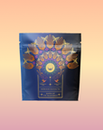 A navy blue tea pouch featuring gold and pink birds, leaves, and a sun design labeled "Queen of the Rainforest: Cacao-Berry Tea," set against a pink-orange gradient—perfect for energizing herbal tea lovers.