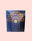 A navy blue tea package features gold and pink birds and leaves in a fan above a sun and crescent moon. The label reads "Queen of the Rainforest: Cacao-Berry Tea," an invigorating herbal blend for an energizing experience.