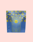 The Queen of Ukraine: Spring Blossom Tea comes in a blue and yellow package with floral patterns, a sun and moon motif, and highlights organic chamomile blossoms and butterfly pea flower on a light pink background.