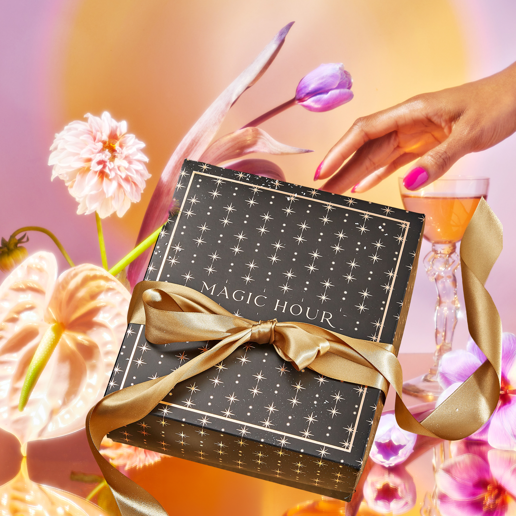 A hand with pink nails reaches for a cocktail near a black Flower Power Mystery Box tied with a gold ribbon, surrounded by pink flowers and glowing colorful light.