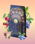 A dark, ornately decorated tea package labeled "Queen of the Rainforest: Cacao-Berry Tea" is surrounded by illustrated fruits, leaves, and blossoms on a pink and yellow gradient background, highlighting this energizing herbal blend.