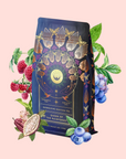A dark, ornate tea bag labeled "Queen of the Rainforest : Cacao-Berry Tea" is surrounded by vivid raspberries, blueberries, cacao pods, and green leaves on a light pink background—reflecting the richness of this chocolate berry tea.