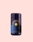 A dark purple tea canister with a vibrant, celestial-themed label reading "Queen of the Rainforest : Cacao-Berry Tea" sits centered on a light pink background. The label shows a sun, stars, and botanicals, evoking Guayusa tea's energizing spirit.