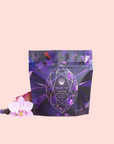 A purple pouch labeled "Amethyst: Strawberry-Passionfruit Gemstone Wellness Tea" stands upright by a pink orchid and lavender sprig, showcasing its organic herbal blend against a pale pink background.