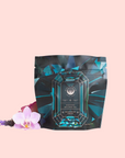 A black and teal geometric tea package labeled "Aquamarine Dream - Soothing Herbal Ayurvedic Adrenal Tonic" rests on a pale pink background, accented by a pink orchid and sprigs of lavender.