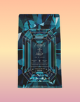 A gemstone-patterned tea bag labeled "Aquamarine Dream - Soothing Herbal Ayurvedic Adrenal Tonic" stands upright against a gradient peach and yellow background, featuring blue geometric shapes and a calming adaptogen sleep blend.