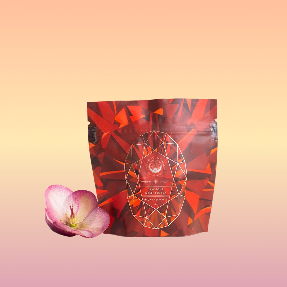 A package of Carnelian: Caffeine-Free Apricot Amaretto Tea with a red geometric design stands upright on a pastel gradient background, beside a single pink orchid blossom.