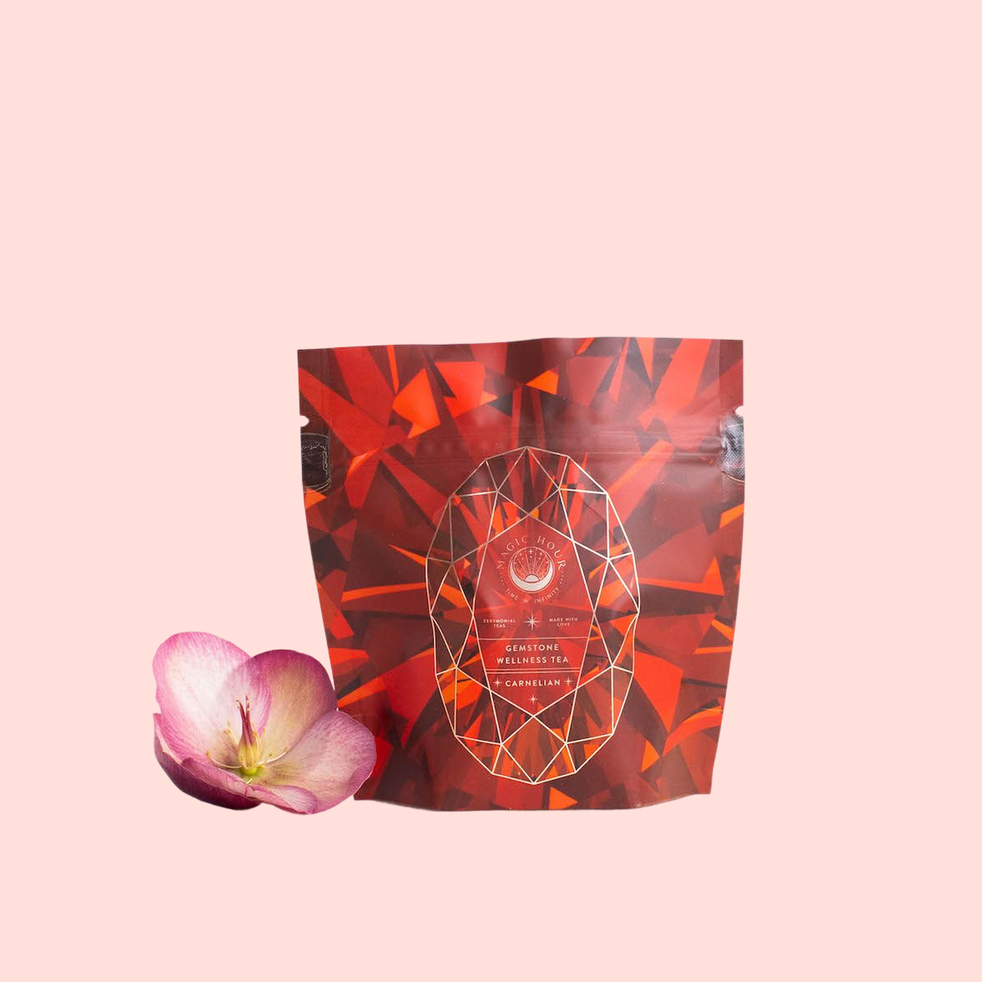 A red and orange geometric-patterned Carnelian: Caffeine-Free Apricot Amaretto Tea package stands upright beside a single pink flower on a pale pink background.