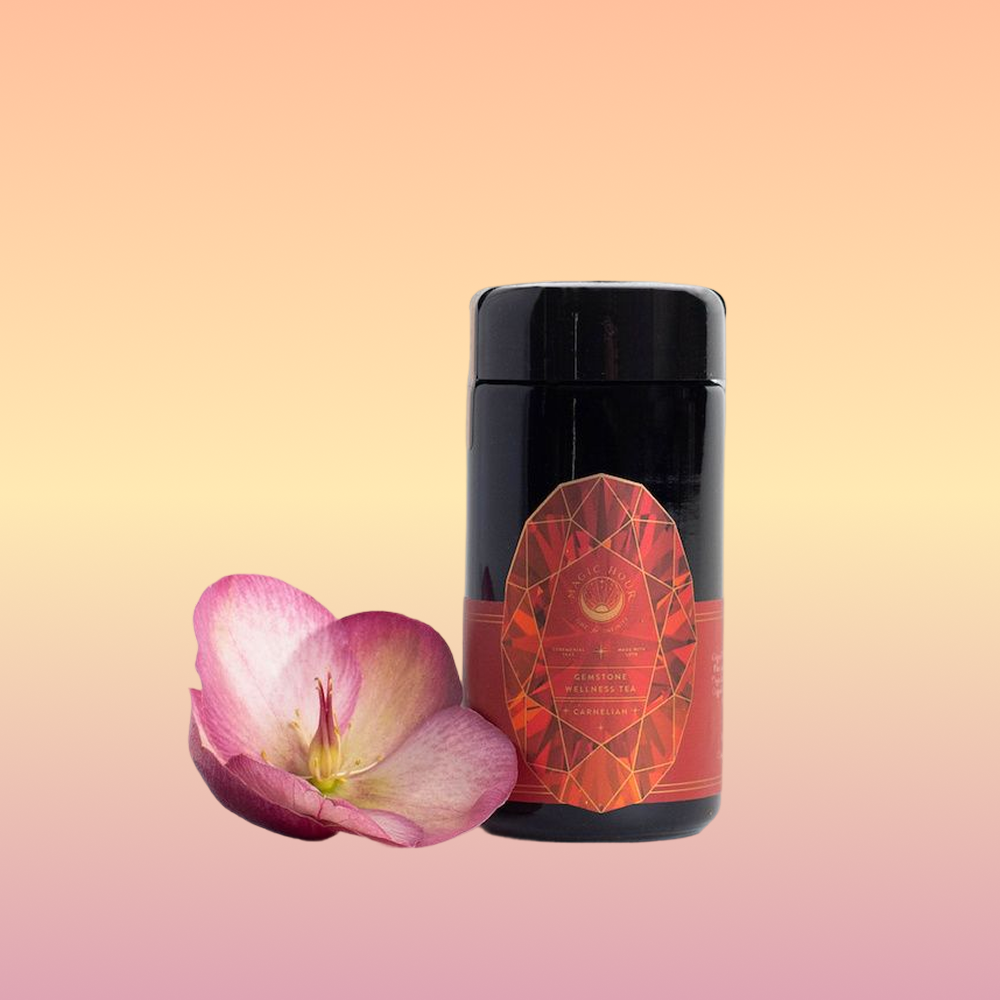 A black jar labeled “Carnelian: Caffeine-Free Apricot Amaretto Tea” stands beside a single pink flower on a soft gradient background of peach, yellow, and pink hues.