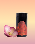 A black jar labeled “Carnelian: Caffeine-Free Apricot Amaretto Tea” stands beside a single pink flower on a soft gradient background of peach, yellow, and pink hues.