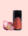 A black cylindrical container with a red geometric label of Carnelian: Caffeine-Free Apricot Amaretto Tea stands beside a pink and yellow flower against a pale pink background—an elegant showcase for this creativity tea.