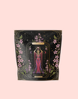 A pale pink package of The Queen of Cups: Hibiscus-Vanilla-Berry Tea for Balance & Heart Health features pink flowers, gold accents, and a woman in a flowing pink dress holding cups, evoking harmony and self-care.