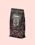 A decorative tea package with floral designs and an illustration of a woman holding a cup. The package reads "Magic Hour" and "The Lovers Hibiscus-Cinnamon-Vanilla Love Potion." A soft pink background adds a romantic touch.