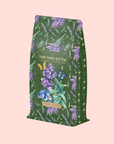 A green and purple Blueberry Lavender Mint: Cosmic Garden Iced Tea package, decorated with blueberries, lavender, butterflies, and stars, stands out against a light pink background. The label highlights this antioxidant-rich iced tea from Magic Hour.