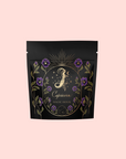 A black stand-up pouch with gold and purple floral design, labeled "Capricorn" and "Magic Hour," showcases Capricorn: Maple Oolong Tea for its metabolism-boosting benefits, set against a pale pink background.
