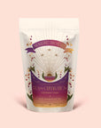 A pouch of Tea for Celebration: Coconut Chai: Rich Spiced Black Tea Blend, adorned with celestial and floral artwork featuring rays, stars, and hands holding a teacup on a light pink background. Infused with blue lotus, this blend is crafted to elevate your senses and awaken the crown chakra.