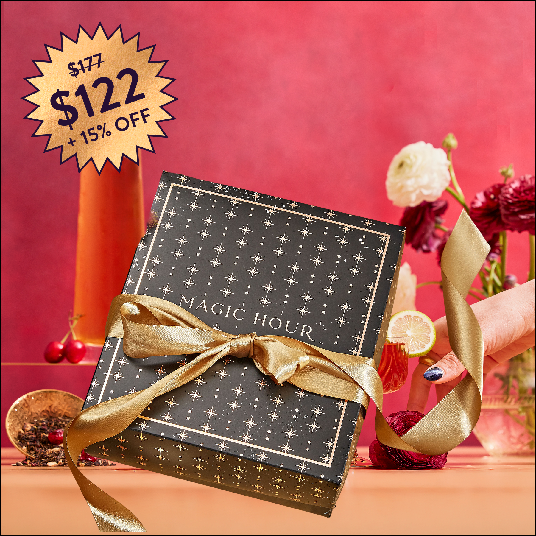 The Memorial Day Mystery Box is held in hand, featuring a black “Magic Hour” gift box with gold stars and ribbon, surrounded by flowers and cocktail. A starburst sign reads “$177 $122 + 15% off.” Ideal for luxury tea lovers!.