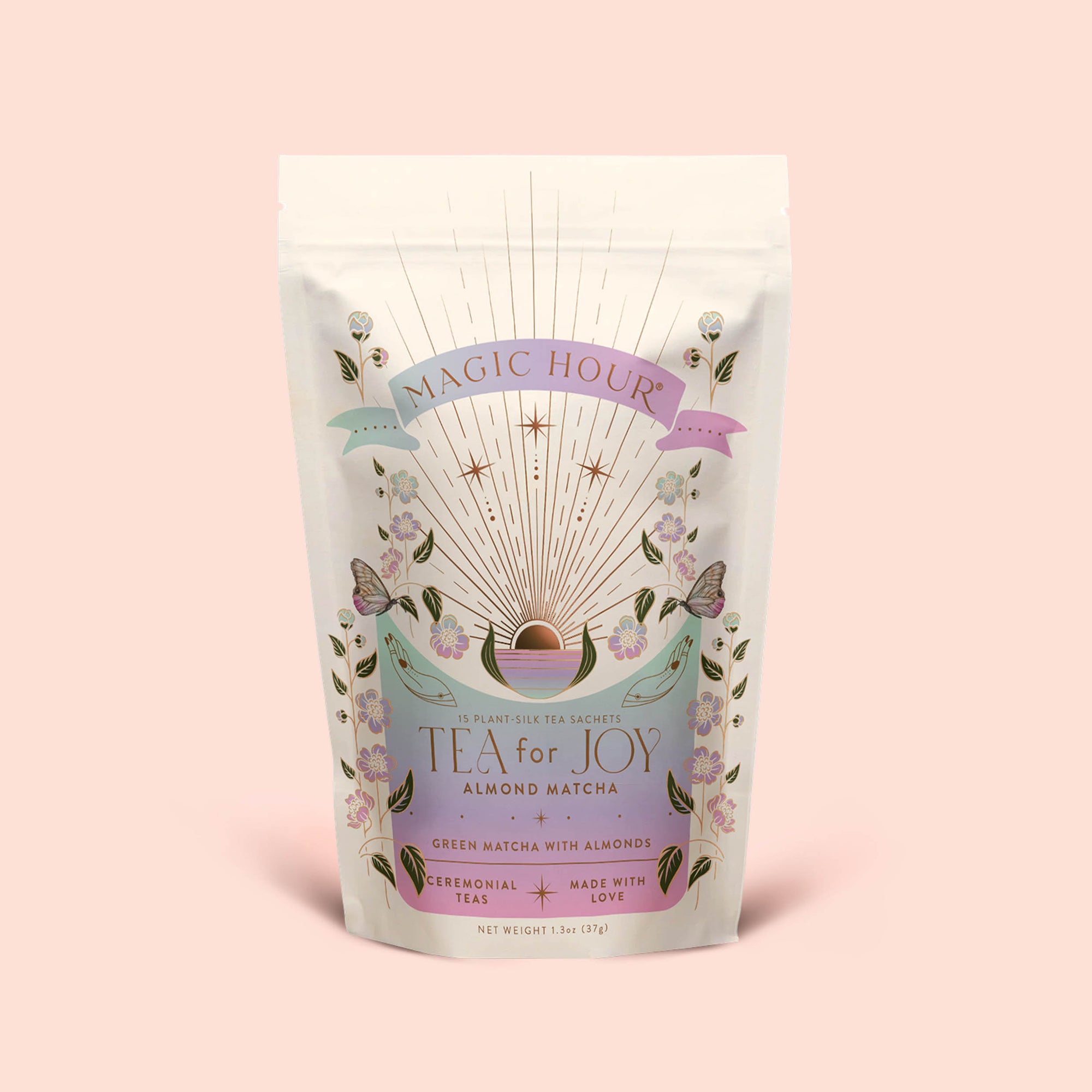 A cream-colored pouch labeled "Tea for Joy: Almond Matcha: Green Matcha Tea with Almonds" adorned with floral and celestial designs rests on a pale pink background. The packaging showcases an antioxidant-rich ceremonial green matcha tea blend featuring almonds and love.