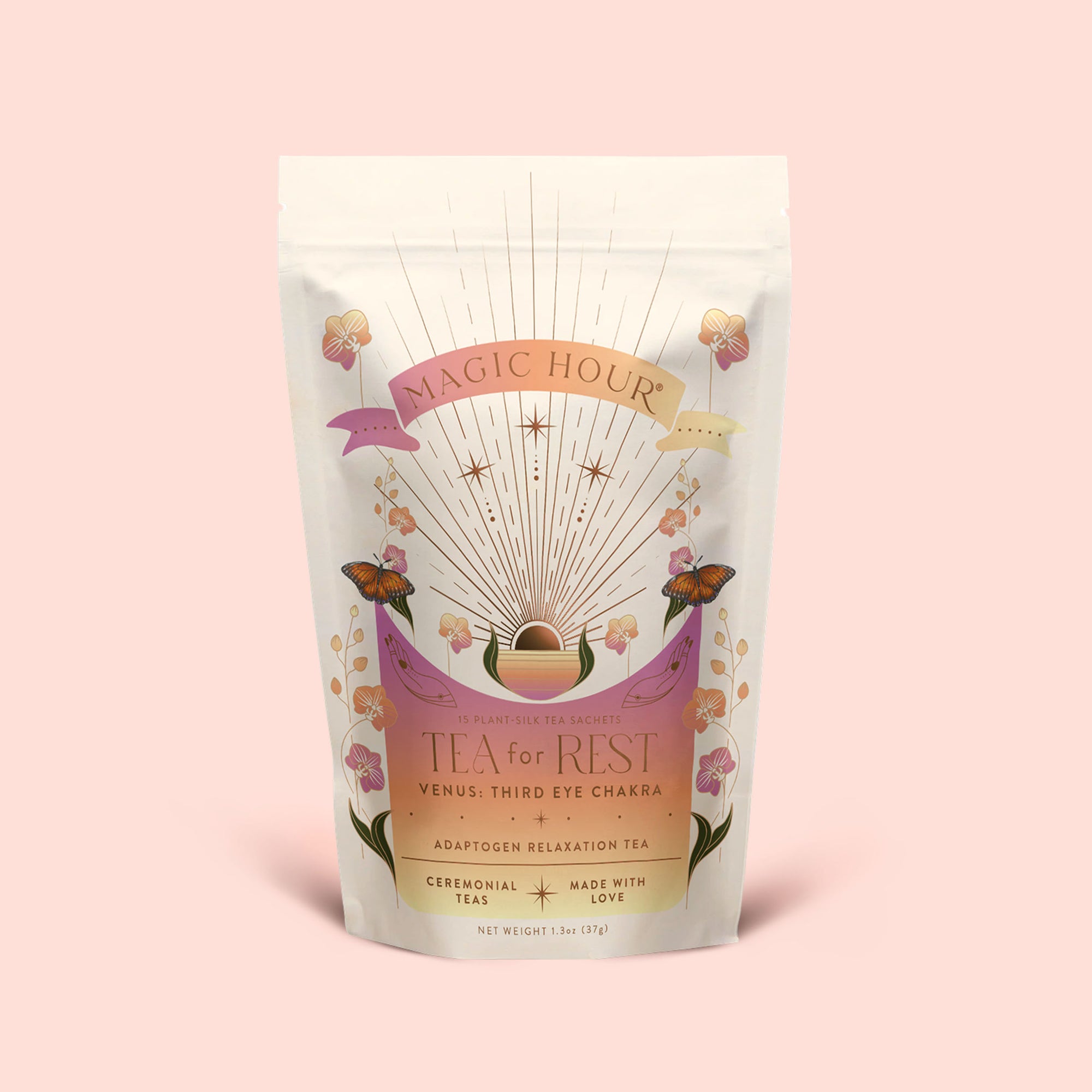 A pastel pouch of Magic Hour’s “Tea for Rest: Venus: Mango Blossom &amp; Nervines Herbal Tea,” adorned with floral and butterfly designs, a celestial motif, and gold accents on a light pink background. This herbal blend features organic chamomile to support relaxation, is labeled as ceremonial, and marked “made with love.”.