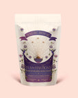 A white and purple Magic Hour tea pouch labeled "Tea for Harmony: Queen of Wellness: Women's Cycle Herbal Blend," featuring floral and celestial designs, with text highlighting it's a women's herbal tea blend with organic chaste berry and raspberry leaf, specially crafted to support the sacral chakra.
