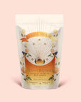 A white pouch of Magic Hour Tea for Cleansing: Queen of the South: Herbal Cafe de Olla features floral designs and sunburst art on a light pink background; this antioxidant-rich, naturally caffeine-free Cocoa Detox Tea is labeled as Root Chakra herbal café de olla.