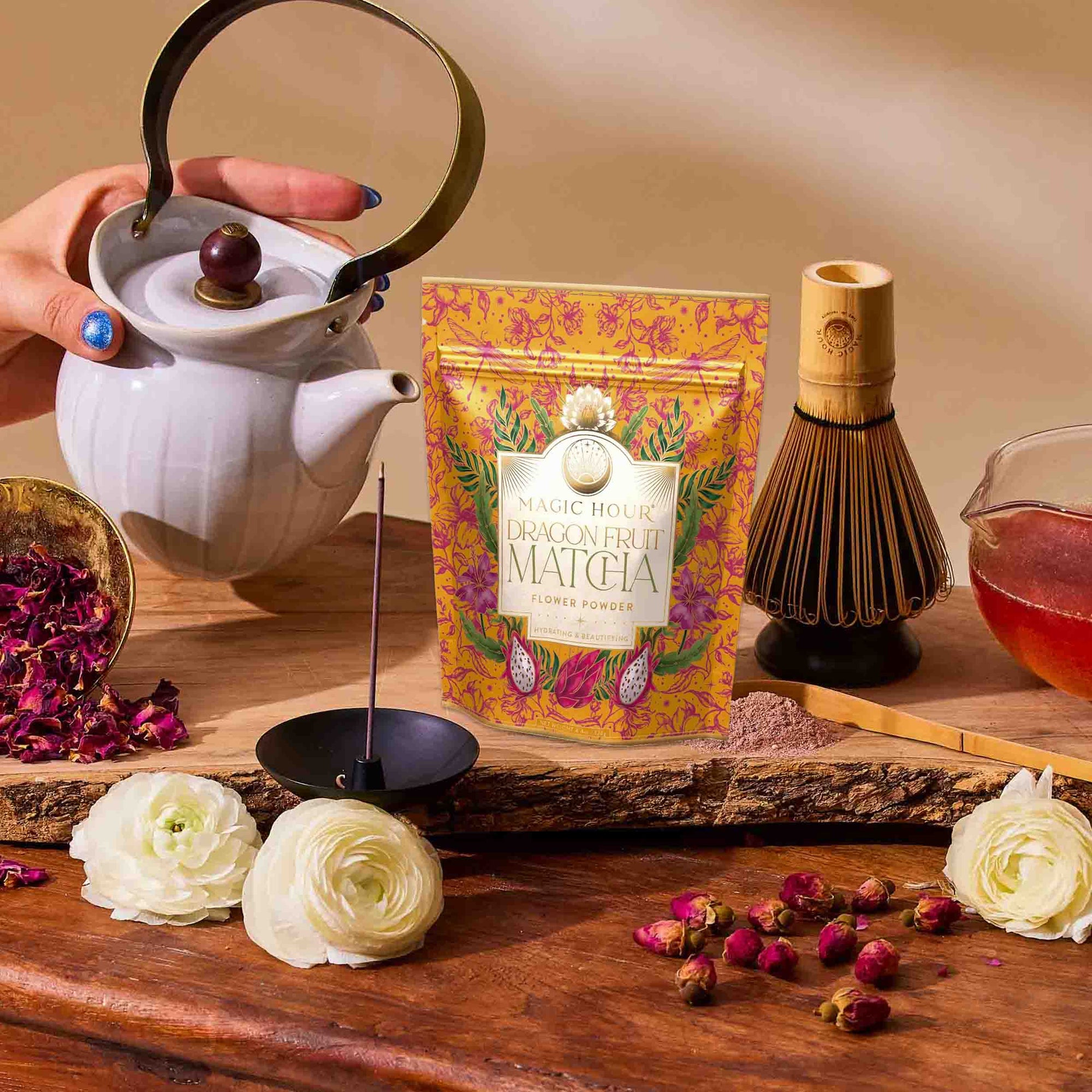 A hand pours luxury tea from a white teapot beside a packet of dragon fruit matcha powder, a bamboo whisk, a bowl of honey, flowers, rose petals, and incense atop a wooden table—capturing the essence of summer refreshments featured in The Best of Summer Poolside Sips Box.