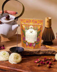 A hand pours luxury tea from a white teapot beside a packet of dragon fruit matcha powder, a bamboo whisk, a bowl of honey, flowers, rose petals, and incense atop a wooden table—capturing the essence of summer refreshments featured in The Best of Summer Poolside Sips Box.