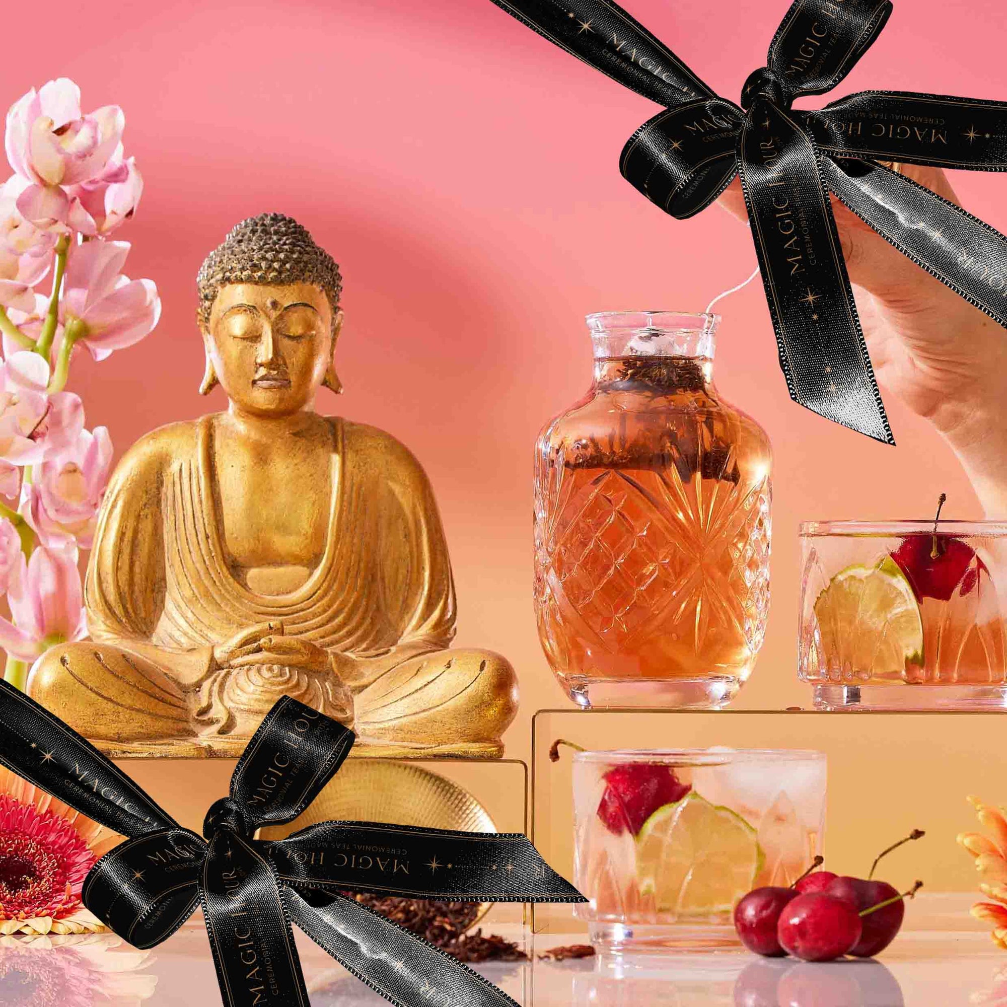 A golden Buddha statue sits beside pink flowers and luxurious tea drinks garnished with cherries, elegantly displayed on glass blocks. Black ribbons with gold text accent the scene, set against a vibrant pink and orange gradient background—creating The Best of Summer Poolside Sips Box, perfect for refreshing summer enjoyment.