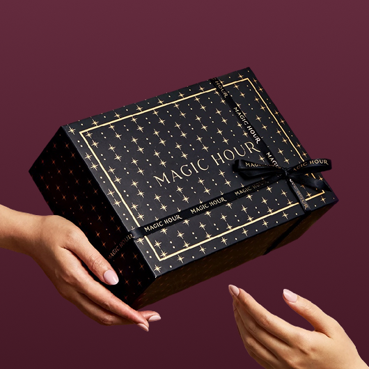 A person hands a Branded Gift Box—black and adorned with gold stars and the words “MAGIC HOUR”—to another person, creating a memorable gifting moment against a rich burgundy background.