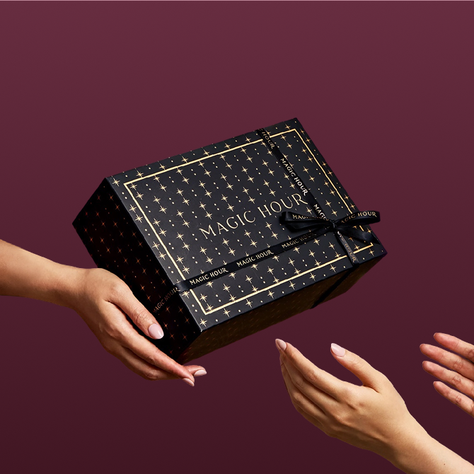 A person hands a Branded Gift Box—black and adorned with gold stars and the words “MAGIC HOUR”—to another person, creating a memorable gifting moment against a rich burgundy background.