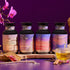 The Balancing Dosha Bundle—four jars of caffeine-free Ayurvedic tea with unique labels—rests on a wooden tray surrounded by an orchid, tea accessories, jewelry, and a glass of tea against a purple backdrop.