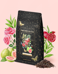 A black tea bag adorned with gold stars and a floral label sits amid roses, guava, pomegranate, leaves, a butterfly, and loose tea leaves on a soft pink background—an elegant presentation that beautifully showcases Guava Pomegranate Black Iced Tea, an organic blend infused with vibrant guava and pomegranate notes.