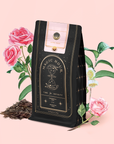 A black bag of Gypsy Rose Black Tea stands before illustrated pink roses and green leaves on a pale pink background, with loose organic black tea leaves scattered at its base.