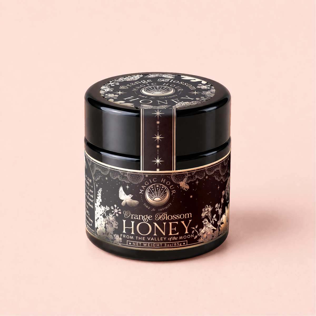 A black glass jar of Orange Blossom Honey: Raw Ojai Honey from the Valley of the Moon, made with organic California honey, featuring floral and bird art on its label, set on a soft pink background.