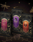 Three ornate coffee bags with Halloween-themed labels—Haunted Mansion, Black Cat Brownie, and Candy Corn—are displayed on a dark, starry background adorned with gold botanical and magical illustrations. The Halloween Bundle captures the spirit of the season with these hand-crafted brews and enchanting tea blends.