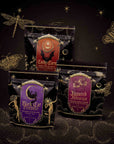 Three decorative tea packages from the Halloween Bundle—“Candy Corn,” “Black Cat Bonfire,” and “Haunted Hibiscus”—are displayed amid gold illustrations of stars, a dragonfly, and a butterfly on a dark, magical-themed background. This Small Batch Magic Halloween Bundle features limited edition selections crafted with hand-blended teas for an enchantingly unique experience.