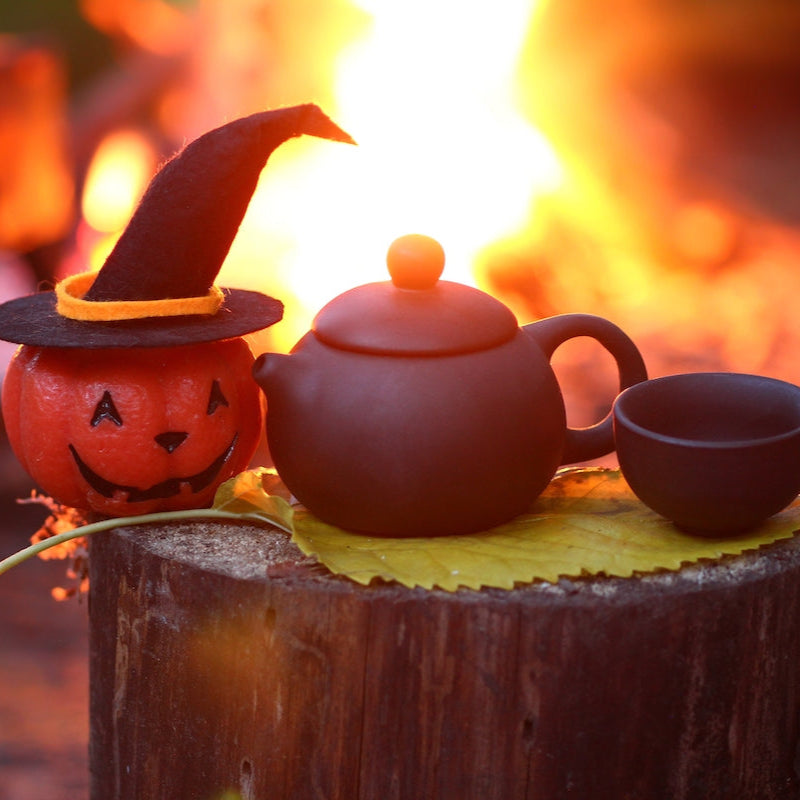 A black teapot and cup rest on a yellow leaf atop a tree stump, beside a pumpkin in a witch hat and a warm fire—perfect for enjoying autumn teas from our Small Batch Halloween Bundle.