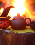 A black teapot and cup rest on a yellow leaf atop a tree stump, beside a pumpkin in a witch hat and a warm fire—perfect for enjoying autumn teas from our Small Batch Halloween Bundle.