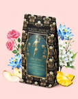 A floral-adorned tea package labeled "Magic Hour" sits amidst illustrated roses, apple slices, pineapple, and delicate white blossoms—evoking the enchanting flavors inside. Set against a pale pink background, the scene invites you to discover delights such as Hathor of Joy: Honey Nectar Herbal Tea nestled within.
