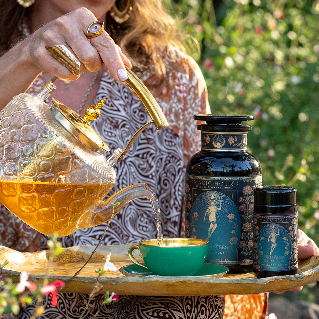 A woman pours Hathor of Joy: Honey Nectar Herbal Tea from a glass teapot into a green teacup on a tray, with tea containers beside her. She wears patterned clothes and gold jewelry, sitting outdoors among flowers and enveloped in the gentle fragrance of honey nectar.