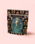 A decorative black tea pouch labeled "Hathor of Joy: Honey Nectar Herbal Tea," featuring an ornate design of a woman with flowers and the text "She Rises—Blue Lotus Honey Nectar Herbal Tea," a caffeine-free blend.