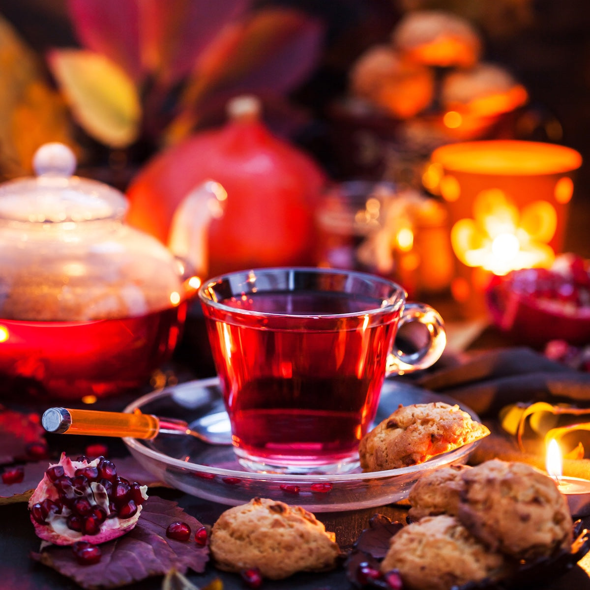 A glass cup of Haunted Hibiscus caffeine-free tea rests on a saucer with cookies, pomegranate seeds, candles, and a teapot nearby. Warm, multicolored lighting sets a cozy autumn or winter mood.